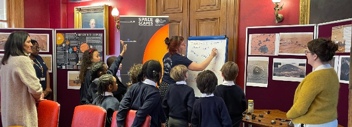 children watching a woman writing on a whiteboard with geological images on display around them